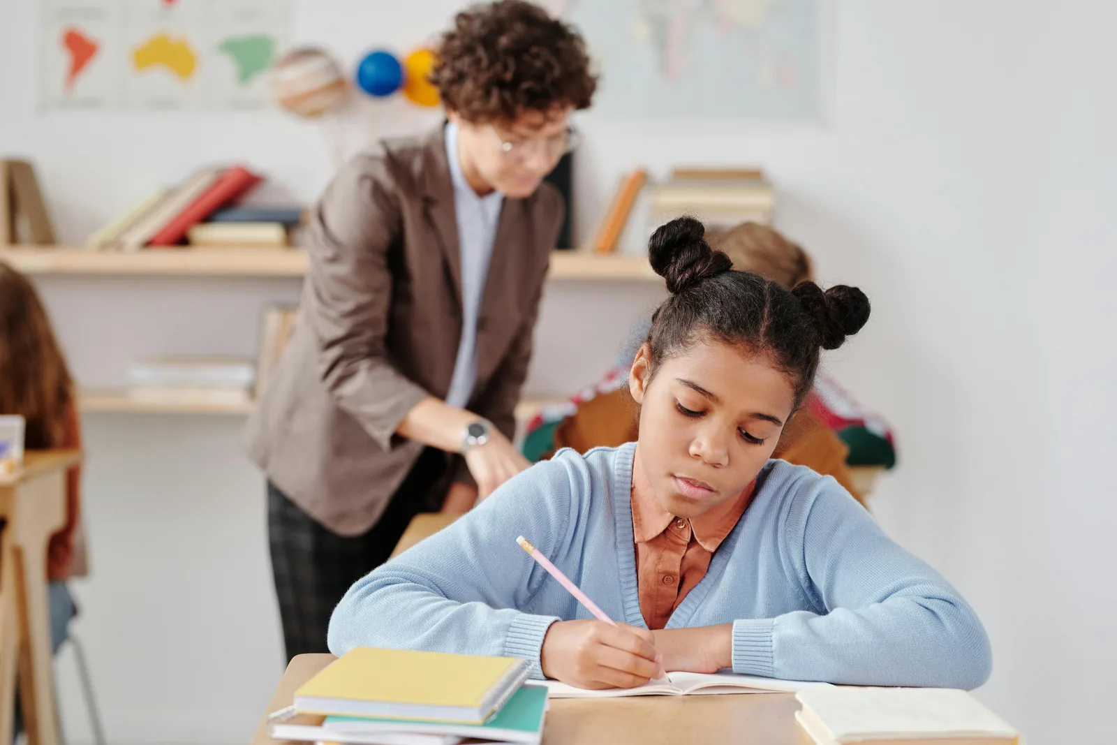 A young student writing at her desk while her teacher works nearby